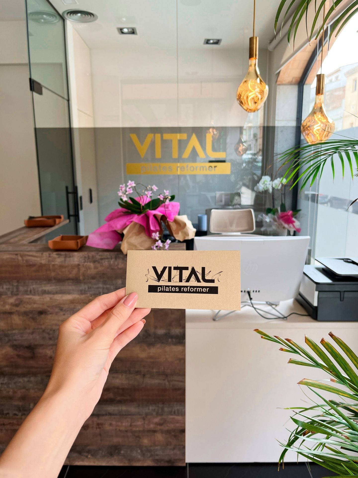 Hand holding card with 'Vital Pilates Reformer' in modern studio with plants and pendant lights.