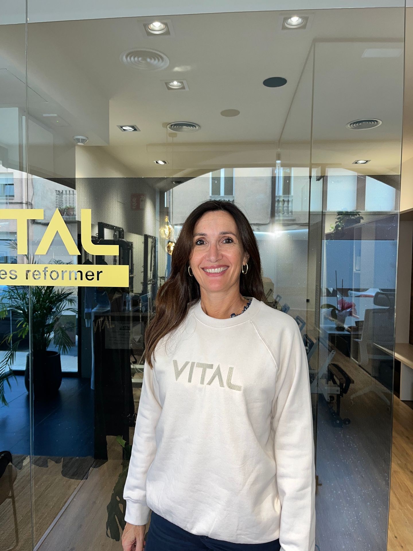 Person smiling in an office lobby with glass doors, wearing a VITAL sweatshirt.